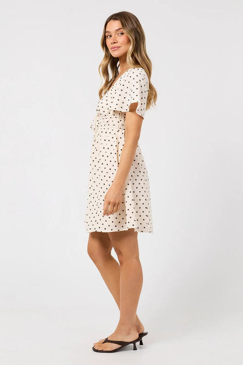 A woman modeling a short-sleeve cream dress with a black polka dot pattern, paired with black heeled sandals, against a plain light gray studio background.