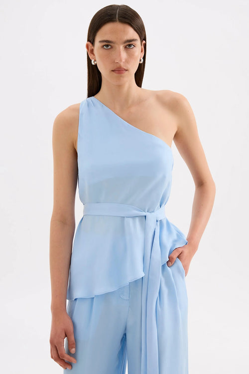 A woman modeling a light blue one-shoulder wrap jumpsuit with matching wide-leg trousers against a plain white studio background.
