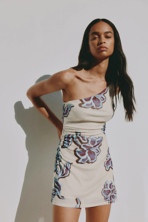 A woman modeling a one-shoulder cream-colored mini dress with a purple and blue floral pattern, standing against a light gray wall with a soft shadow.