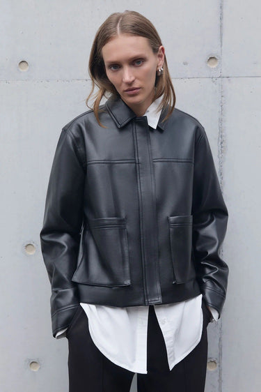 Person wearing a black leather jacket over a white shirt against a gray concrete wall.