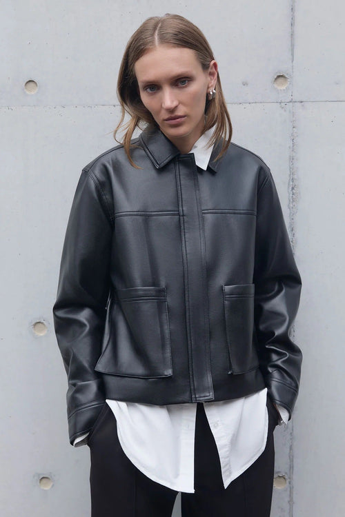 Person wearing a black leather jacket over a white shirt against a gray concrete wall.