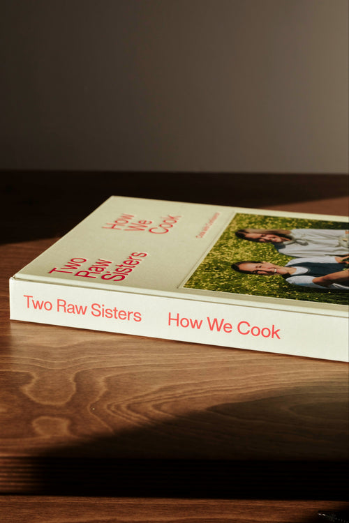 The book 'How We Cook: Two Raw Sisters' resting on a dark wooden table under warm, directional sunlight, casting a soft shadow on a dark gray background.