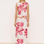 A woman stands against a plain off-white wall wearing a sleeveless white maxi dress with a large pink floral print and a matching waist tie belt. The floor is a light beige woven texture.