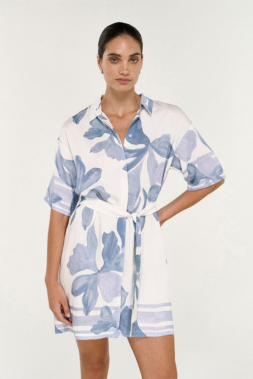 A woman modeling a white short-sleeve shirt dress with a blue floral print and a matching tie belt, set against a plain light gray studio background.