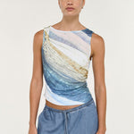 A woman modeling a sleeveless crop top with an abstract blue, white, and beige wave-like print, paired with light blue denim pants, against a plain light gray studio background.