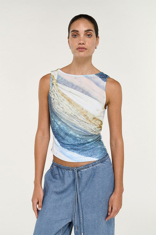 A woman modeling a sleeveless crop top with an abstract blue, white, and beige wave-like print, paired with light blue denim pants, against a plain light gray studio background.