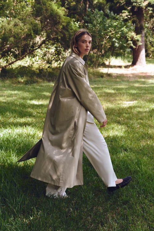Person wearing a beige coat and white pants walking in a park.