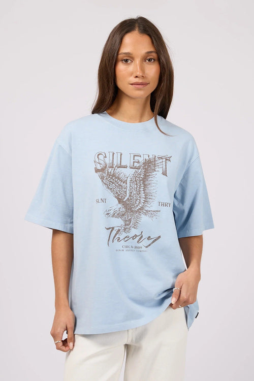 A woman modeling a light blue oversized graphic t-shirt with a distressed eagle design and the text 'SILENT Theory' on the front, paired with off-white pants against a light gray studio background.