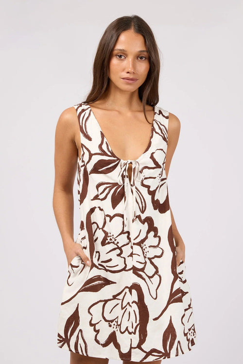 A woman modeling a sleeveless, scoop-neck dress with a brown and cream floral print, standing against a solid light gray studio background.
