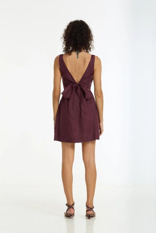 A woman viewed from the back wearing a sleeveless deep burgundy mini dress with a large tie-back bow, paired with brown strappy sandals, against a plain white studio background.