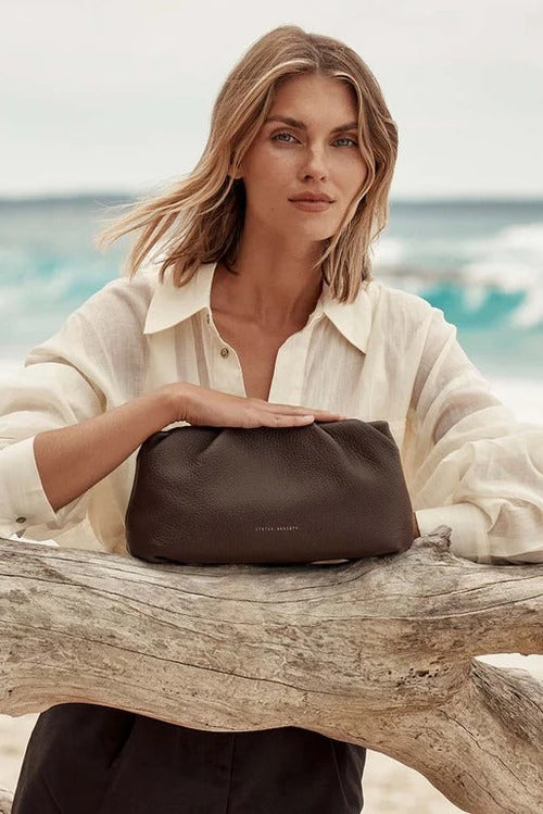 A woman in a cream button-up shirt sits on a driftwood log at the beach, holding a dark brown leather pouch bag. The background shows a soft-focus ocean scene.
