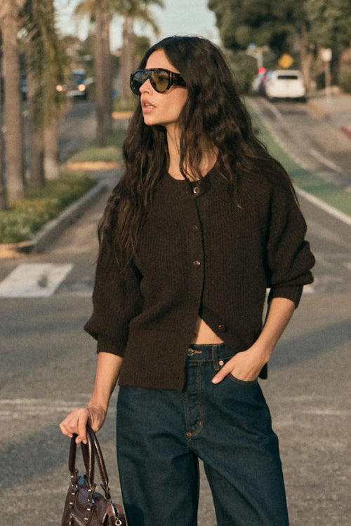 Woman in a brown cardigan and sunglasses standing on a street.