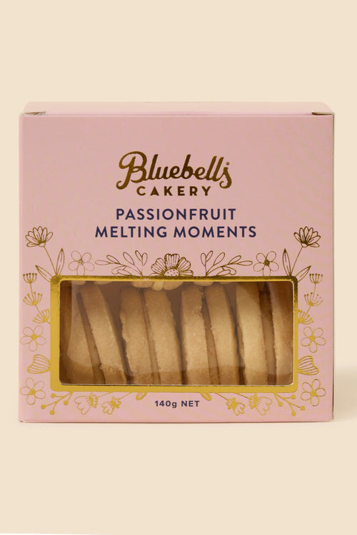 A pink box of Bluebell's Cakery Passionfruit Melting Moments, featuring a clear window that displays the golden-brown biscuit slices inside, set against a solid cream background.