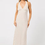 A woman modeling a sleeveless halter-neck maxi dress with a white base and small black polka dots, paired with black flat sandals against a plain light gray studio background.