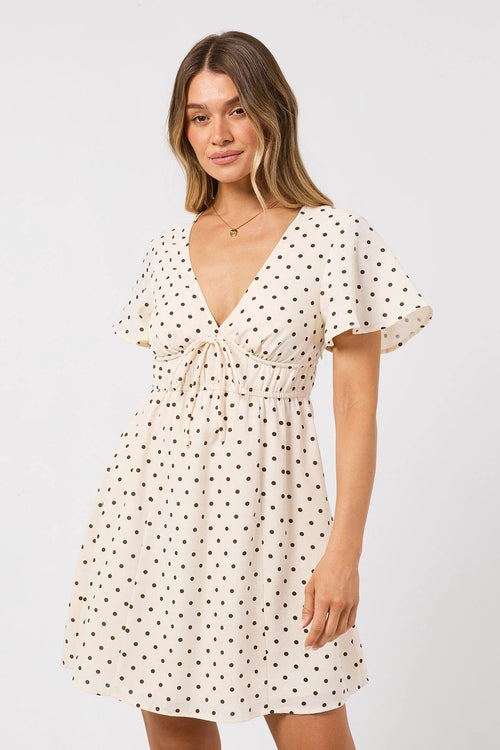 A woman modeling a short-sleeve cream dress with a black polka dot pattern, a V-neckline, and a drawstring waist, set against a plain light gray background.
