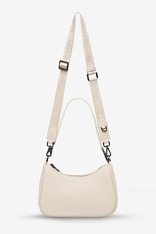 A light beige leather crescent-shaped shoulder bag with a matching branded webbed strap, displayed on a clean, light gray background.