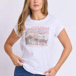 A woman modeling a white short-sleeve graphic t-shirt with a vintage-style 'French Riviera' print on the front, paired with a blue denim skirt, against a plain white studio background.