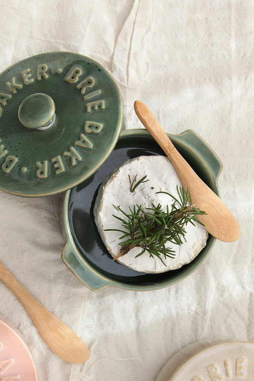 A ceramic Brie cheese warmer dish with its lid off, showing a round wheel of Brie cheese topped with a sprig of rosemary inside. A wooden spoon rests on the dish, all set on a textured cream linen background.