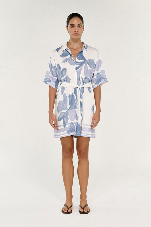 A woman modeling a short-sleeve white shirt dress with a blue floral print, featuring a button-down front, collar, and self-tie belt, set against a plain light background.