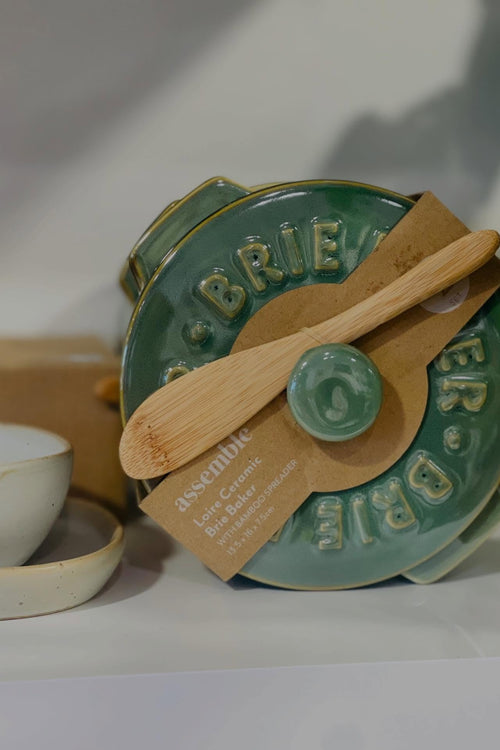 A green ceramic Brie cheese mold with a bamboo spatula and a cardboard tag reading 'assemble', photographed on a light surface with a softly blurred background.