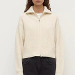 Woman wearing a cream ribbed sweater over a white shirt with black pants on a white background