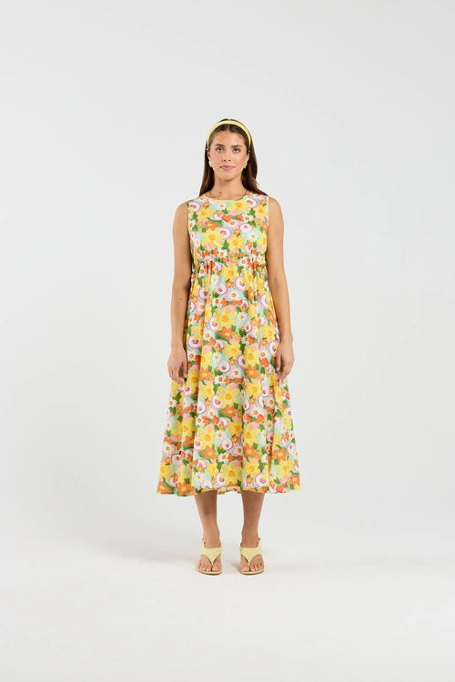 A woman stands against a plain light gray studio background wearing a sleeveless midi dress with a bright floral print in yellow, white, pink, and green. She is also wearing a matching headband and tan heeled sandals.