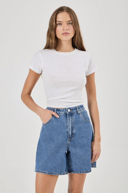A woman modeling a white short-sleeve crewneck t-shirt and high-waisted blue denim shorts against a light gray studio background.
