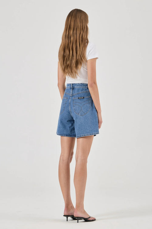 A woman viewed from behind wearing light-wash blue denim shorts, a white t-shirt, and black heeled sandals against a plain light gray studio background.