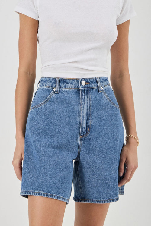 A person wearing light-to-medium wash blue denim shorts with a white t-shirt, shown from the chest down against a plain light background.