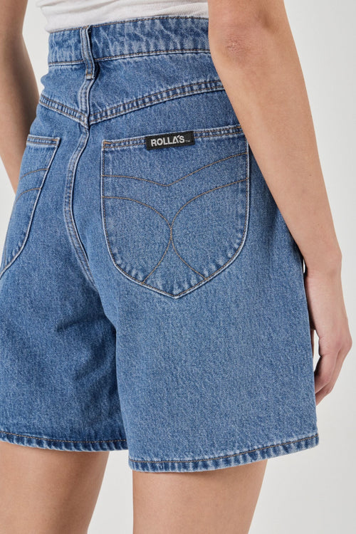 Close-up back view of a person wearing blue denim shorts with a visible ROLLO'S brand patch on the back pocket, against a plain light background.