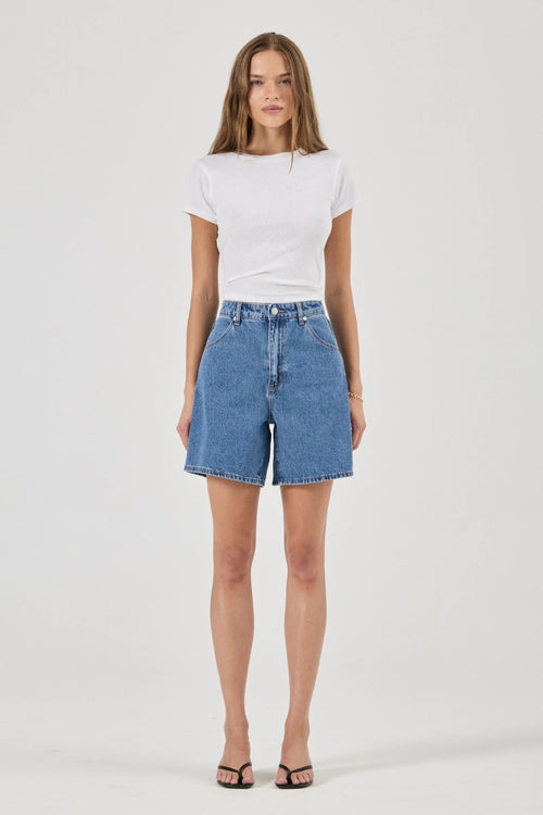 A woman modeling a white short-sleeve crewneck t-shirt and high-waisted light blue denim shorts against a plain light gray studio background.