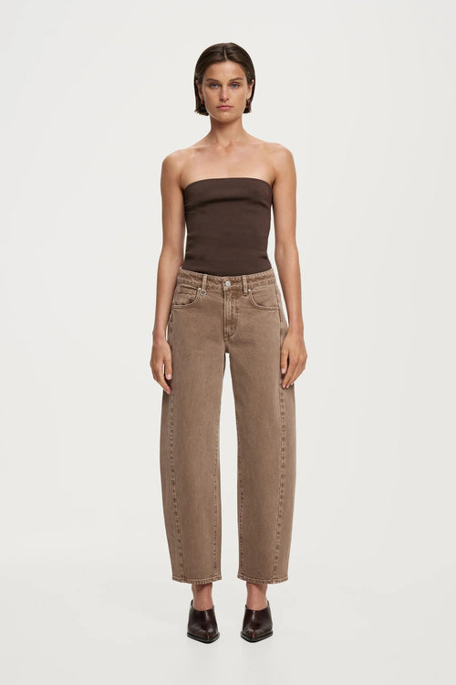 A model stands against a light gray studio background wearing a dark brown strapless tube top and wide-leg cropped jeans in a warm taupe-brown color with decorative studs down the outer seams, paired with dark brown pointed shoes.