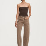 A model stands against a light gray studio background wearing a dark brown strapless tube top, beige straight-leg jeans with contrast stitching, and black pointed-toe heels.