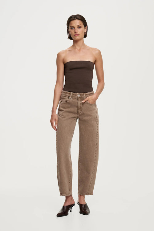 A model stands against a light gray studio background wearing a dark brown strapless tube top, beige straight-leg jeans with contrast stitching, and black pointed-toe heels.