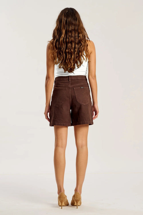 A woman viewed from behind wearing brown corduroy shorts, a white ribbed tank top, and tan high heels against a plain light gray studio background.
