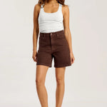 A woman modeling a pair of dark brown high-waisted shorts with a white ribbed tank top and beige heels, standing against a plain white studio background.