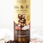 A tall cylindrical chocolate gift box labeled 'The Remarkable Chocolate Make Me Kit' stands on a light marble surface with scattered chocolate pieces, marshmallows, and red candy flakes around its base. The box is cream and dark brown with printed chocolate imagery and product text.