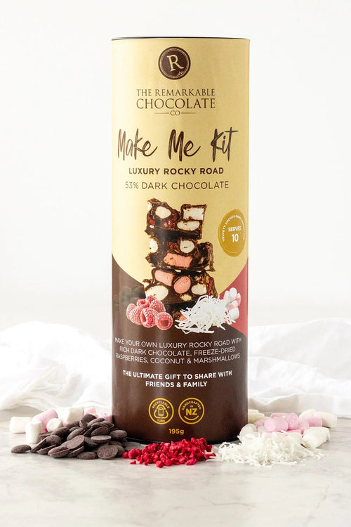 A tall cylindrical chocolate gift box labeled 'The Remarkable Chocolate Make Me Kit' stands on a light marble surface with scattered chocolate pieces, marshmallows, and red candy flakes around its base. The box is cream and dark brown with printed chocolate imagery and product text.