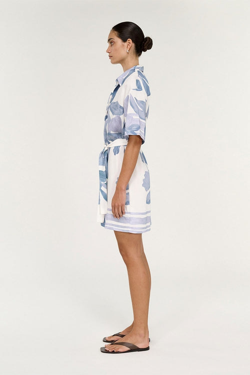 A woman stands in profile wearing a short white shirt dress with a blue watercolor floral print and a matching tie belt, paired with dark brown flip-flop sandals, against a plain light gray studio background.