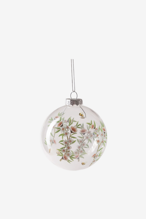 Clear glass bauble, decorated with a delicate print of white manuka blossoms, soft green foliage, and buzzing bees.