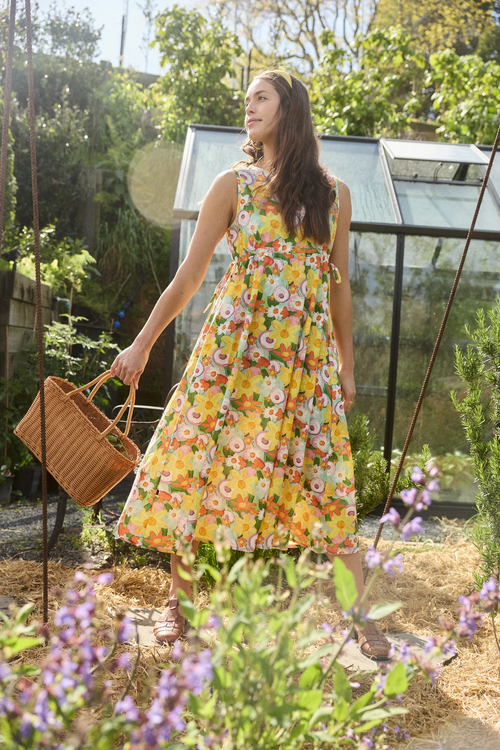 A woman stands in a sunlit garden wearing a sleeveless yellow floral dress and holding a woven basket. A greenhouse is visible in the background among green plants.