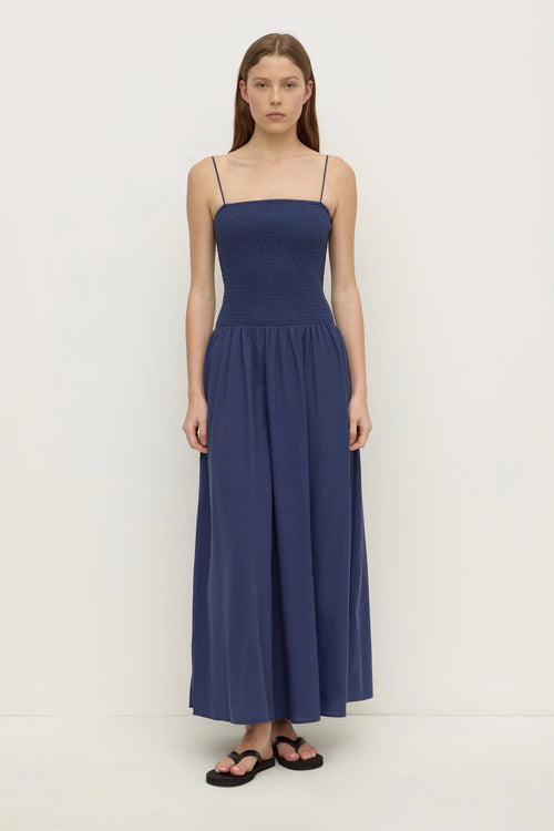 A woman modeling a navy blue maxi dress with thin spaghetti straps, a smocked bodice, and a flowing skirt, paired with black sandals against a plain white studio background.