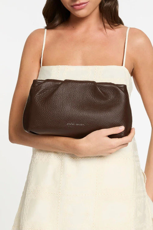 A woman wearing a cream-colored spaghetti strap dress holds a dark brown pebbled leather clutch bag with a small gold logo that reads 'STATUS ANXIETY' on a plain white background.