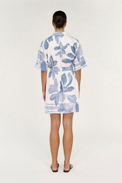 A woman viewed from behind wearing a short white wrap-style dress with a blue floral print and a matching tie belt, standing against a plain light gray studio background.