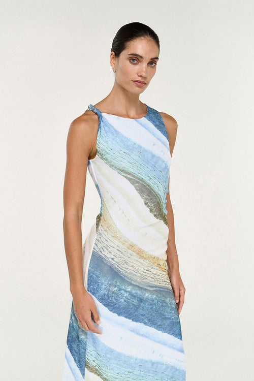 A woman modeling a sleeveless, one-shoulder dress with an abstract beach-inspired print in shades of blue, white, beige, and gray, set against a plain off-white studio background.