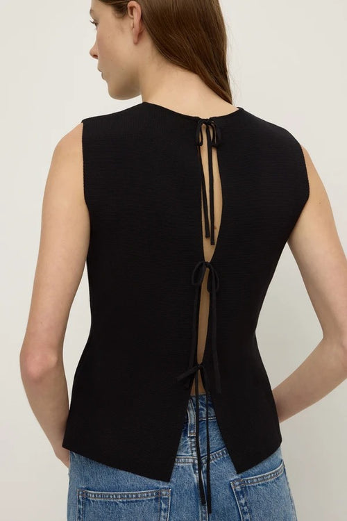 A woman shown from the back wearing a sleeveless black top with a tie-back closure and blue jeans, photographed against a plain light background.