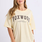 A woman modeling an oversized pale yellow Foxwood graphic t-shirt paired with lavender shorts and white sneakers, standing against a plain white studio background.