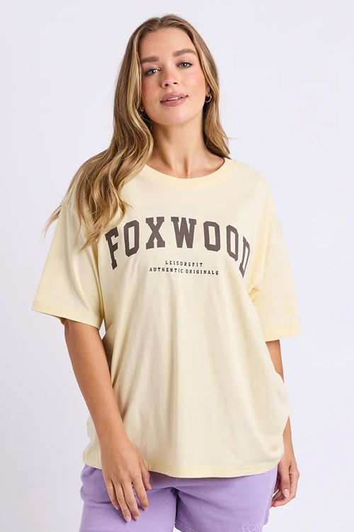 A woman modeling an oversized pale yellow Foxwood graphic t-shirt paired with lavender shorts and white sneakers, standing against a plain white studio background.