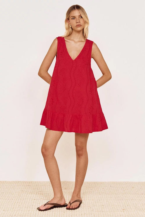 A woman modeling a sleeveless red mini dress with a V-neckline and ruffled hem, paired with dark flip-flop sandals, set against a plain off-white wall and light woven floor.