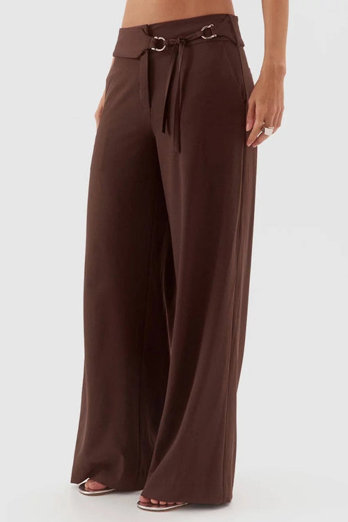 A person wearing high-waisted, wide-leg brown pants with a drawstring waist and decorative metal rings, shown against a plain light gray studio background.